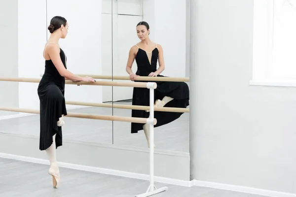 Graceful ballerina in black dress training at barre near mirrors — Stock Photo