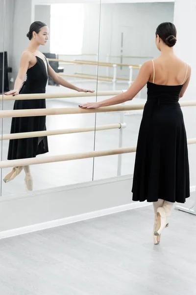 Visão de comprimento total da bailarina em vestido preto ensaiando perto de espelhos em estúdio — Fotografia de Stock