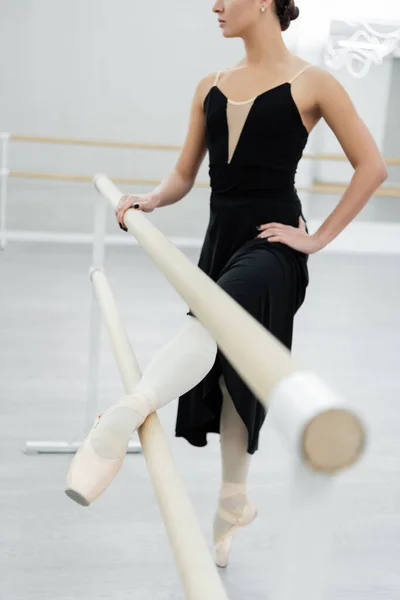 Partial view of ballerina in black dress exercising with hand on hip at barre — Stock Photo