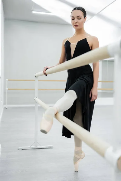 Ballerine en robe noire s'étirant à la barre dans la salle de danse — Photo de stock