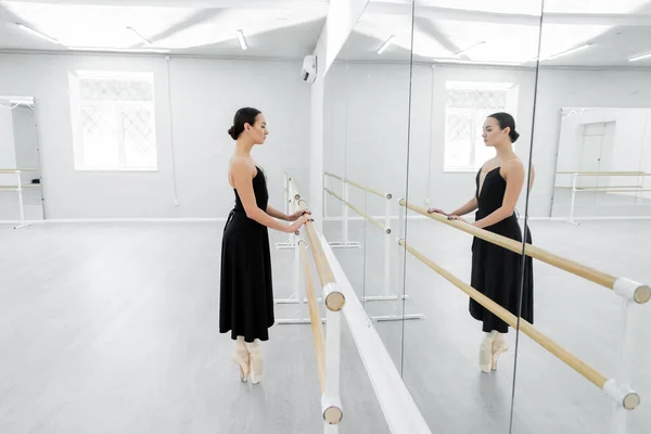 Vue latérale du danseur de ballet en robe noire debout sur les orteils près de la barre en studio — Photo de stock