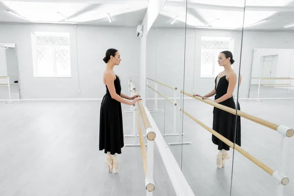 Side view of ballet dancer in black dress standing on toes near barre in studio — Stock Photo