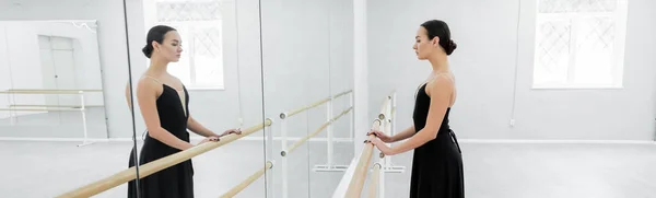 Brunette woman in black dress standing at barre near mirrors in ballet studio, banner — Stock Photo