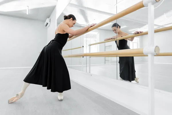 Молодая балерина в черном платье тренируется в баре возле зеркал — стоковое фото