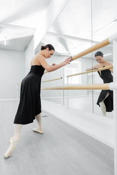 Dançarina de balé em vestido preto exercitando no barre perto de espelhos — Fotografia de Stock