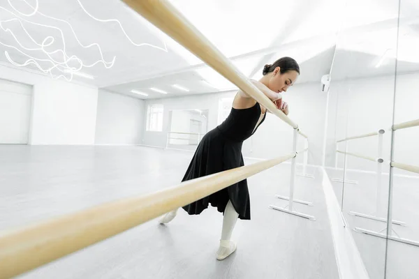 Mujer delgada en vestido negro entrenamiento en barra en estudio de ballet - foto de stock