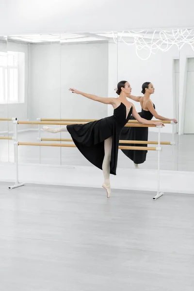 Vue pleine longueur de ballerine mince en robe noire répétant près de barre et miroirs — Photo de stock