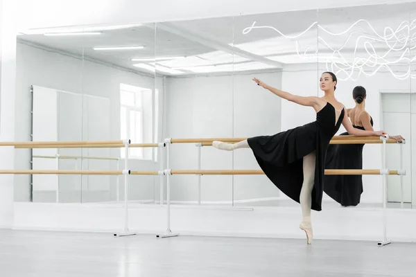 Jovem bailarina em vestido preto ensaiando no barre perto de espelhos — Fotografia de Stock