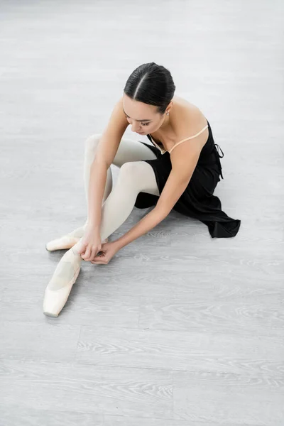 Young ballerina adjusting pointe shoe while sitting on floor in studio — Stock Photo