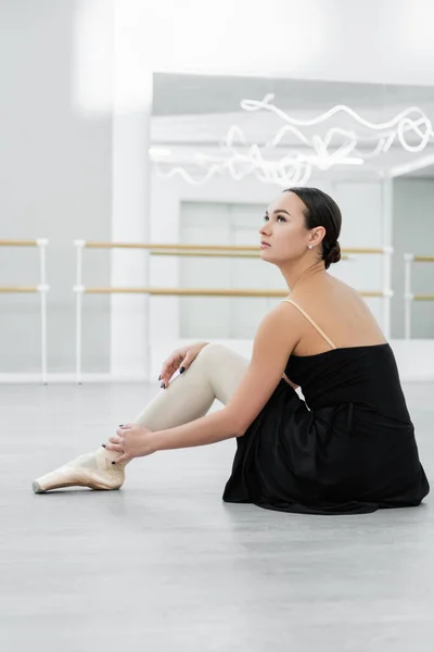 Jeune ballerine en robe noire assise sur le sol en studio et regardant ailleurs — Photo de stock