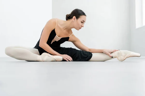 Joven bailarina en vestido negro estirándose en el suelo en el estudio de ballet - foto de stock