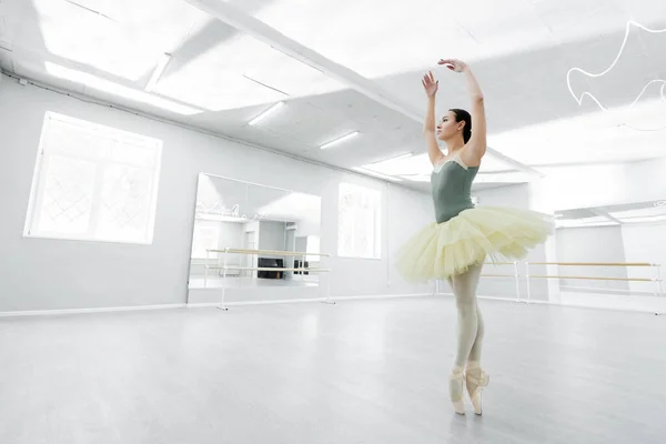 Full length view of ballerina in tutu dancing with raised hands in spacious studio — Stock Photo