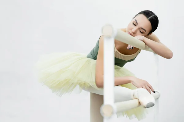 Bailarina com os olhos fechados esticando a perna no barre durante o exercício em estúdio — Fotografia de Stock