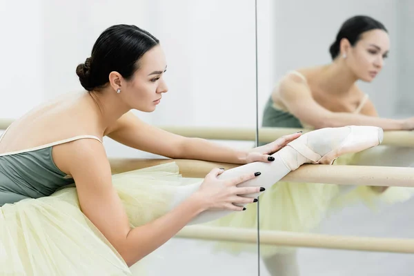 Брюнетка балетная танцовщица растягивает ногу в баре возле зеркал в студии — стоковое фото