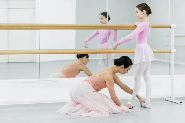 Profesora de ballet tocando pies de niña entrenando en barra cerca de espejos - foto de stock