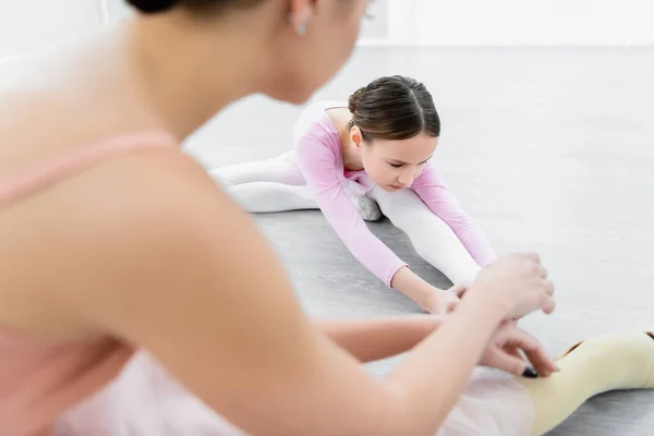 Mädchen streckt sich in Ballettschule neben verschwommenem Tanzlehrer auf dem Boden — Stockfoto