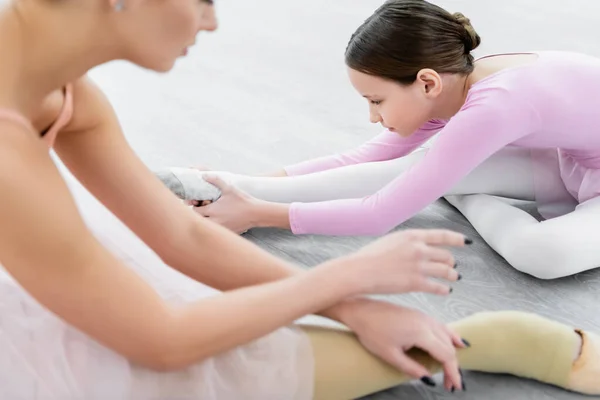 Chica estiramiento en piso en ballet escuela cerca borrosa profesor - foto de stock
