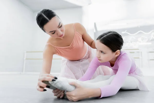 Preteen girl looking at dance teacher while stretching on floor in ballet school — Stock Photo