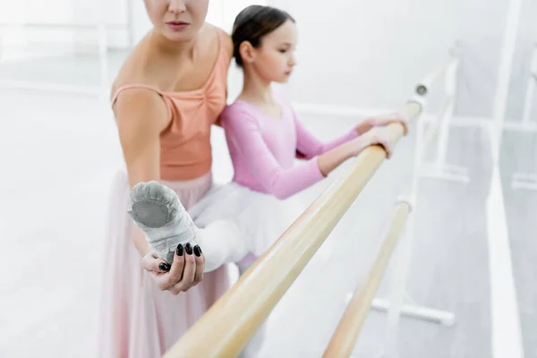 Ballet teacher helping girl stretching at barre in dance studio, blurred background — Stock Photo