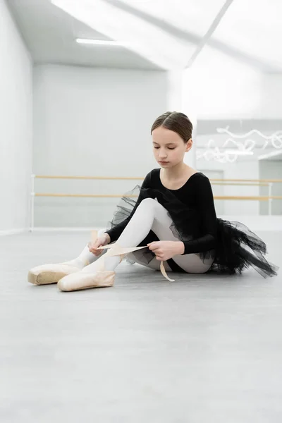 Kind im schwarzen Ballettkostüm bindet Spitzenschuh auf dem Boden im Tanzsaal — Stockfoto