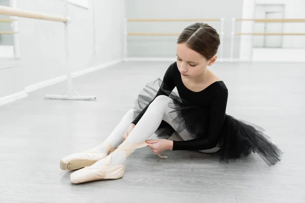 Girl sitting on floor in dancing hall and tying pointe shoe — Stock Photo