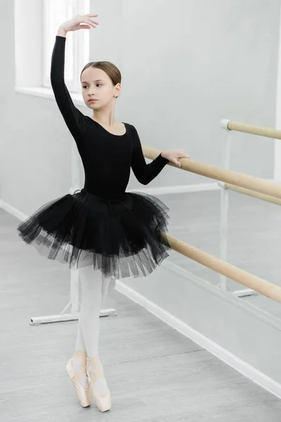 Chica delgada en tutú negro de pie en el dedo del pie mientras se entrena en la barra en el estudio de baile - foto de stock