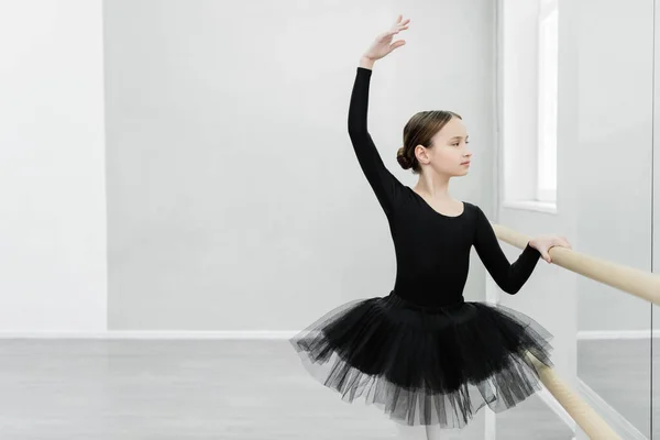 Preteen girl in black tutu looking in mirror while training at barre in ballet studio — Stock Photo