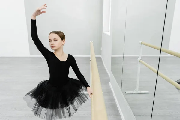 Graceful girl in black tutu training with raised hand at barre near mirrors — Stock Photo