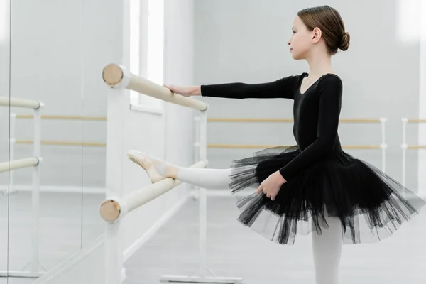Side view of kid in black tutu stretching at barre in dancing hall — Stock Photo
