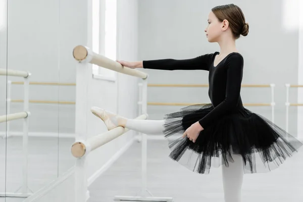 Seitenansicht eines Kindes in schwarzem Tutu, das sich an der Barre im Tanzsaal dehnt — Stockfoto