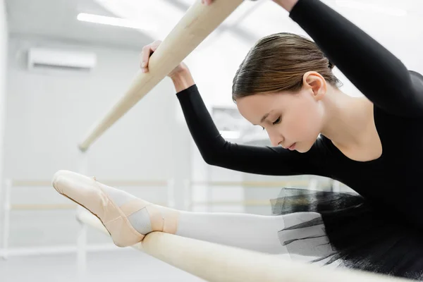 Enfant en costume noir étirant la jambe à la barre en studio de ballet — Photo de stock