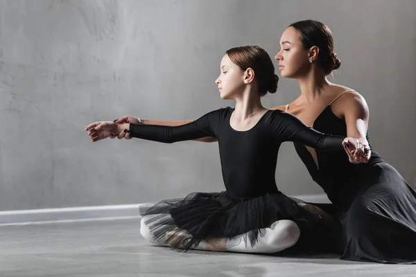 Ребенок в черной пачке сидит со скрещенными ногами рядом с учителем танцев во время урока балета — стоковое фото
