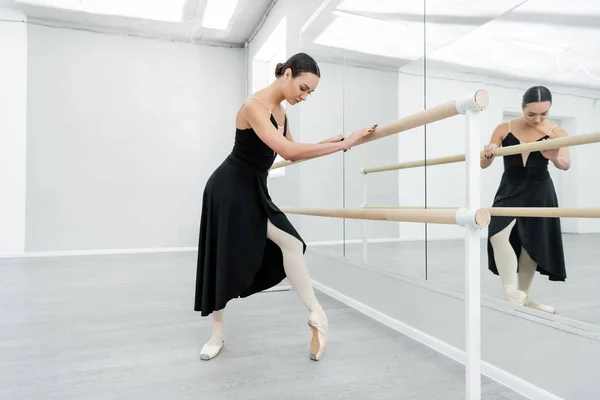 Vista completa de la bailarina en vestido negro y zapatos de punta de entrenamiento en la barra cerca de los espejos - foto de stock