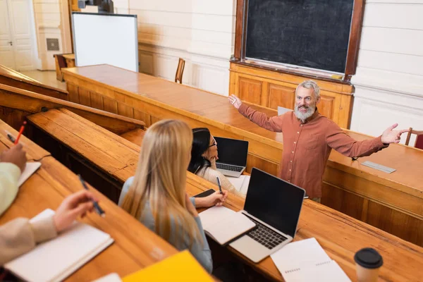 Professora sorrindo segurando giz perto de estudantes multiétnicos no auditório universitário — Fotografia de Stock