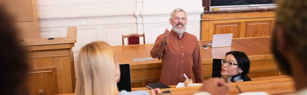 Smiling teacher pointing with hand at blurred african american student in university, banner — Stock Photo