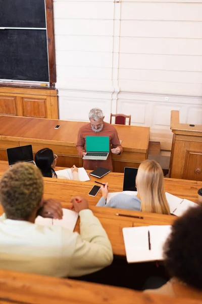 Mature teacher holding laptop with green screen near multiethnic students in university — Stock Photo