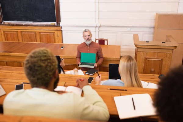 Enseignant tenant un ordinateur portable avec écran vert près des étudiants multiethniques dans l'auditorium de l'université — Photo de stock