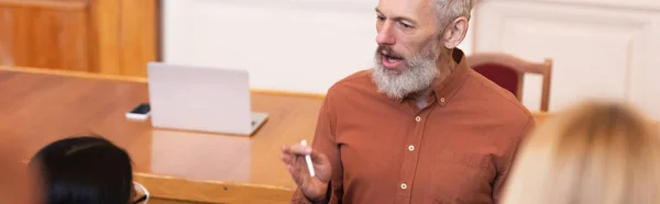 Middle aged professor holding chalk near blurred students in university, banner — Stock Photo