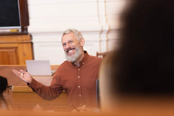 Усміхнений вчитель тримає дошку біля розмитих студентів в аудиторії університету — стокове фото
