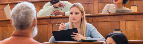 Interracial students writing on notebooks near blurred teacher in university, banner — Stock Photo