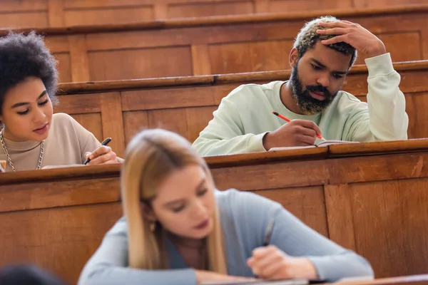 African american student writing on notebook near friends in university auditorium — Stock Photo