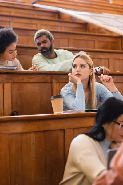 Pensive student sitting near notebook, coffee to go and interracial friends in university — Stock Photo