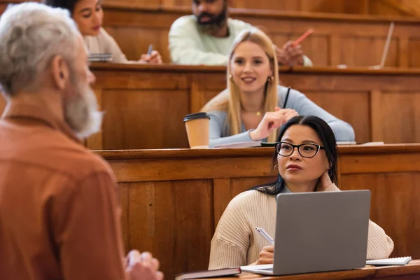 Asiatischer Student sieht verschwommenen Lehrer in der Nähe von Laptop und Notebooks an der Universität — Stockfoto