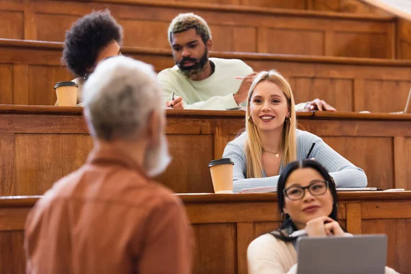 Des étudiants souriants et multiethniques regardent le professeur près du café et des cahiers à l'université — Photo de stock