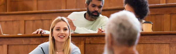 Smiling student looking at blurred teacher near african american friends in university, banner — Stock Photo