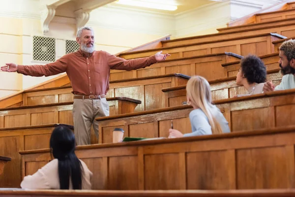 Cheerful mature professor holding chalk near multiethnic students in university — Stock Photo