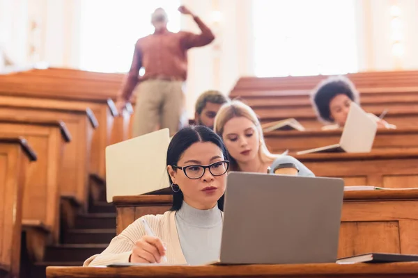 Estudante asiático escrevendo no notebook perto de laptop e amigos multiétnicos borrados na universidade — Fotografia de Stock