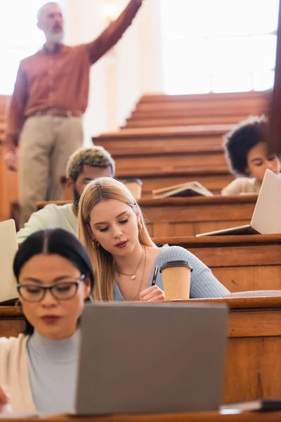 Young student writing near coffee to go, multiethnic friends and teacher in university — Stock Photo