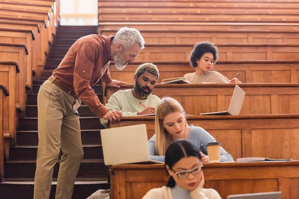 Maturo insegnante in piedi vicino a studenti multietnici scrivere su notebook vicino computer portatili in università — Foto stock