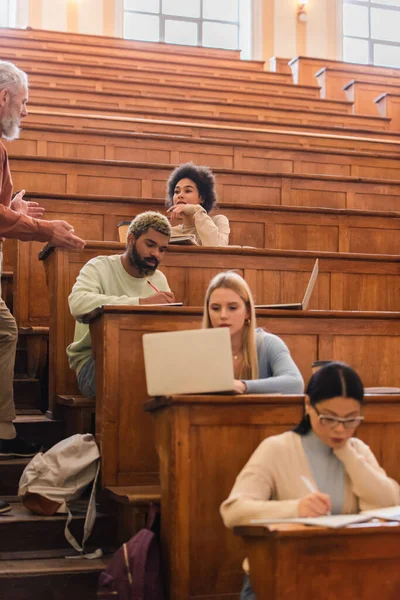 Estudiante afroamericano mirando al profesor cerca de amigos interracial escribiendo en cuadernos en la universidad — Stock Photo